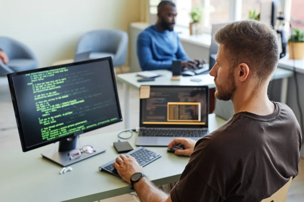 Man is engaged with a computer, while another man sits next to him, both involved in rigorous testing and bug fixes