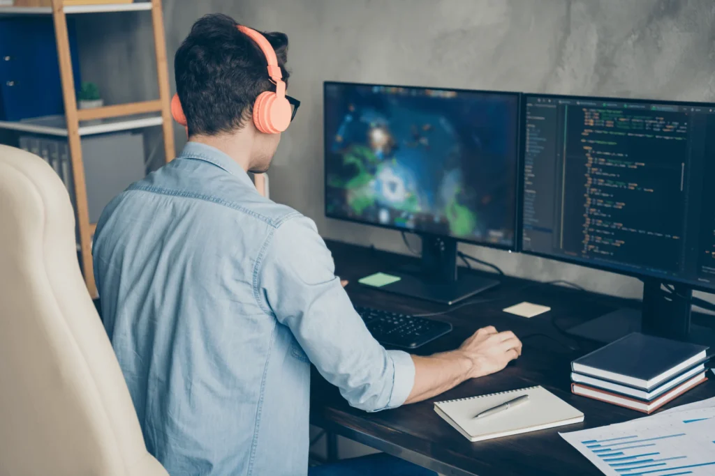 Man wearing headphones works at a desk with two computer monitors, immersed in game development tasks